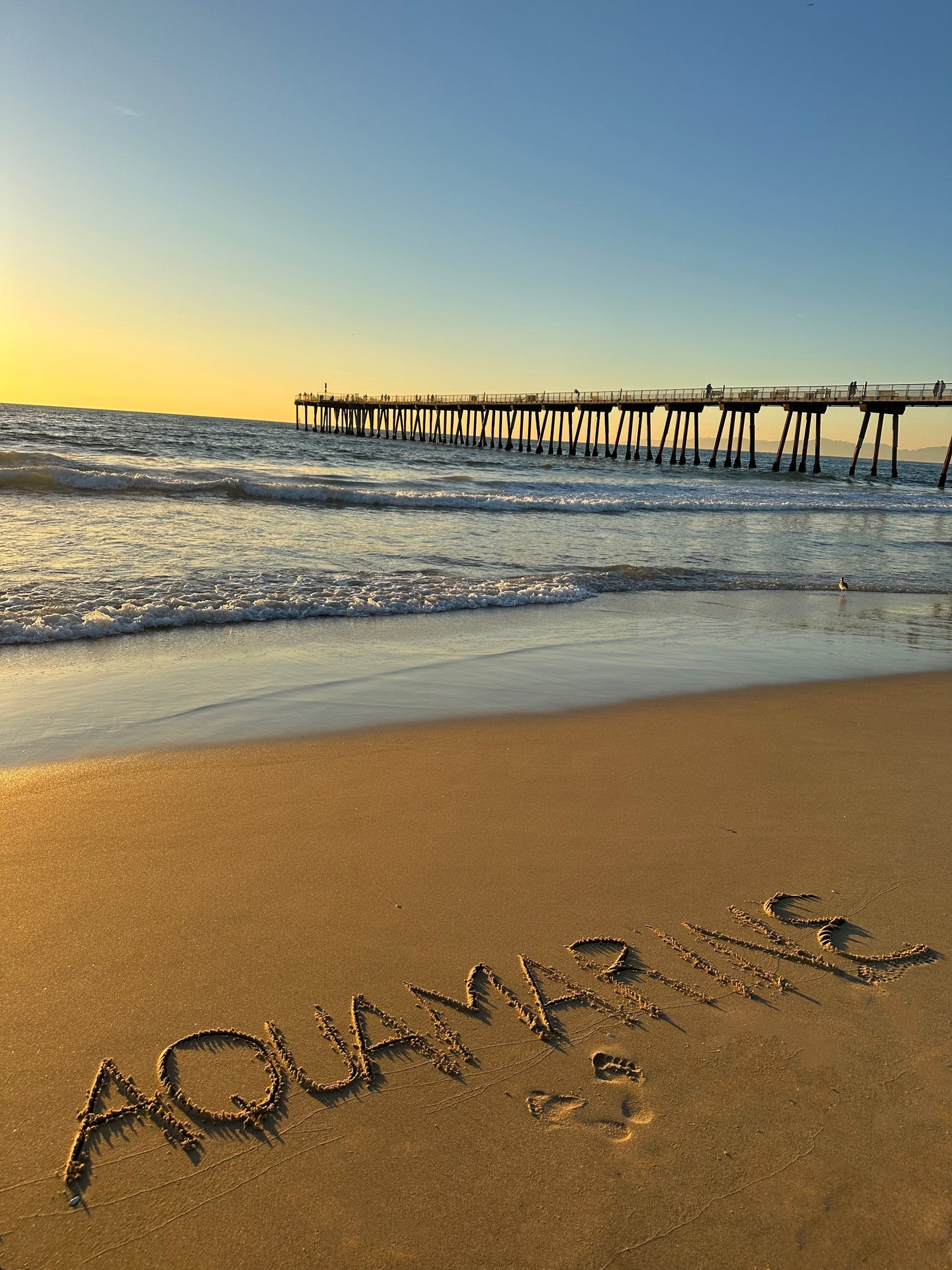 The word 'Aquamarine' elegantly written in the sand on a beach, symbolizing the color, art, and design inspirations behind Aquamarine Design Studio, reflecting the sea's tranquility, serenity, and the deep personal significance it holds for the artist.